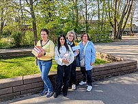 Eine Gruppe von vier Frauen steht vor einer kleinen Mauer im Grünen.