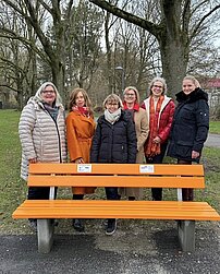 Eine Gruppe von sechs Frauen steht hinter einer orangefarbenen Bank und lächelt in die Kamera.