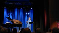 Eine in blau gekleidete Frau steht neben einem Flügel auf der Bühne und spricht in ein Mikrofon.
