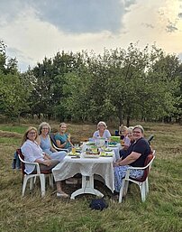 Eine Gruppe von Frauen sitzt um einen Tisch mit weißer Decke auf einer Wiese.